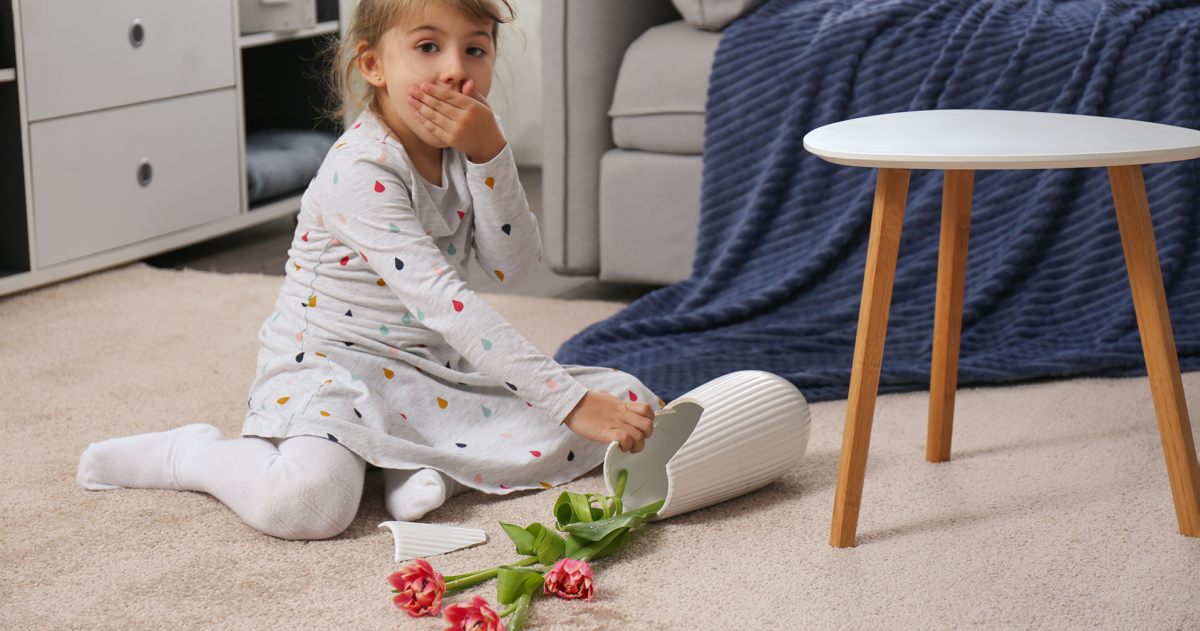 Een klein meisje schrikt en heeft haar hand voor haar mond omdat de vaas met bloemen gevallen en gebroken is
